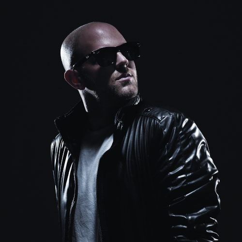 Randi Seidman DJ is looking slightly over his left shoulder wearing dark sunglasses and in a leather jacket. He has subtle lighting showing the reflection shine off his jacket with a solid black background