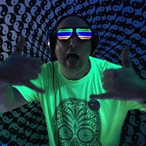 DJ Term has rainbow striped sunglasses on. They are glowing UV along with his neon yellow t shirt which has a sugar skull on it. He has his tongue sticking out with both hands held out front of him in a fist with the thumb and little fingers poking out in a cool gesture